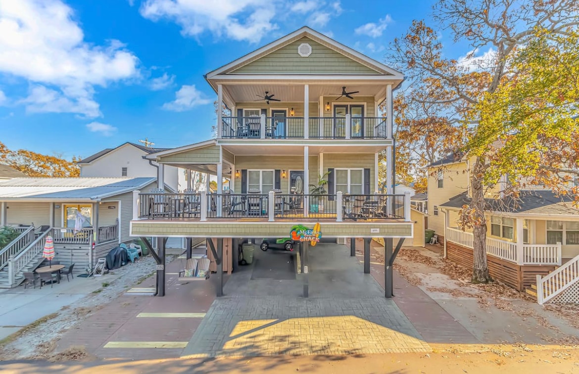 A custom beach house is showcased from the exterior, featuring a three-level structure with multiple balconies. The ground level is partially enclosed, providing space for parking. Surrounding trees and blue skies create a pleasant outdoor environment, inviting guests to explore the nearby area.
