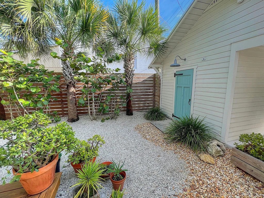 A serene outdoor space is surrounded by lush green palm trees and various potted plants. Soft gravel covers the ground, with pathways leading to a green door, enhancing the natural ambiance of the area.
