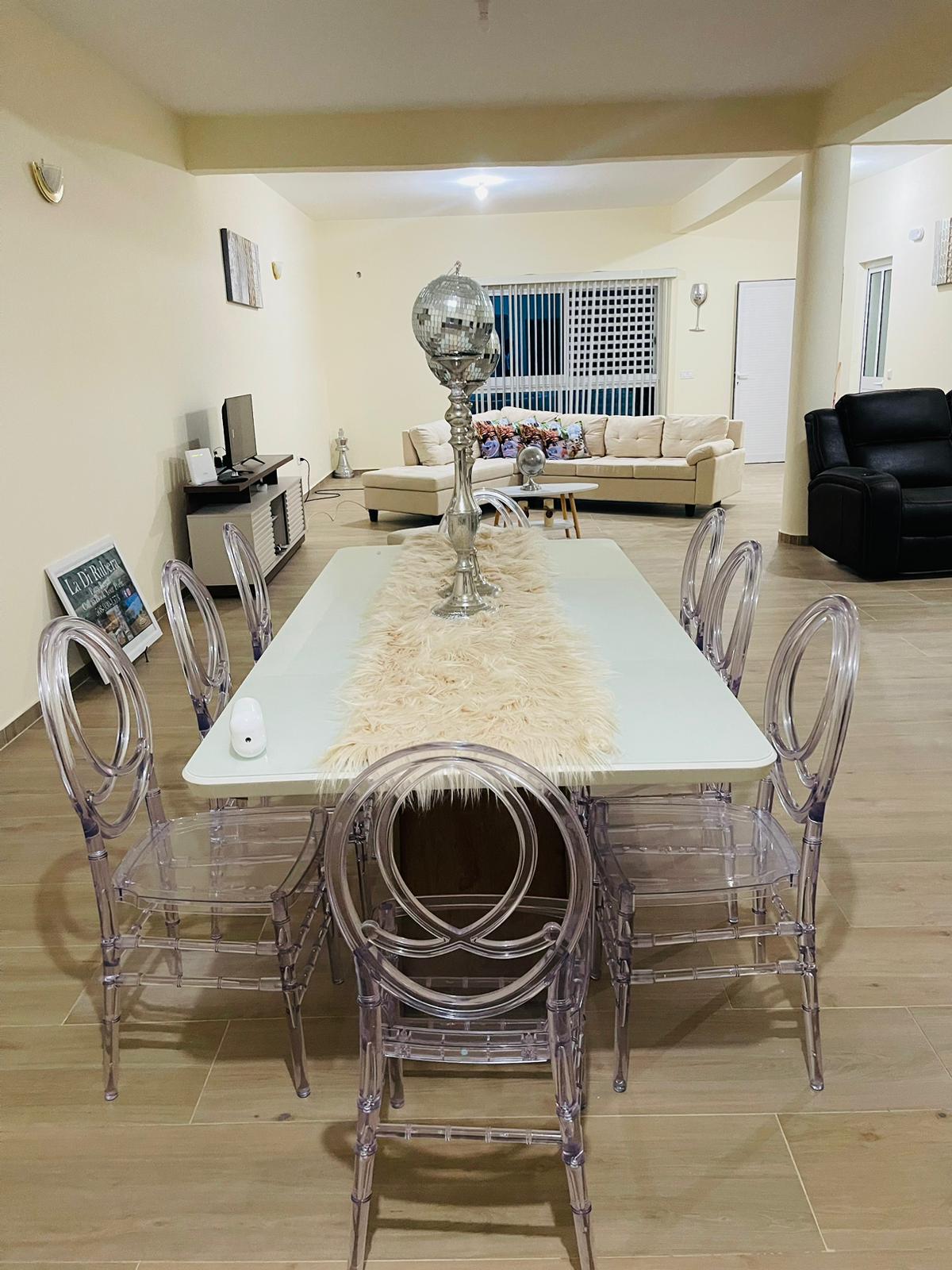 A long dining table is set with a light-colored table runner and is surrounded by six transparent chairs. A decorative lamp stands centrally on the table. In the background, a cozy living area features a light-colored sofa and a dark armchair.