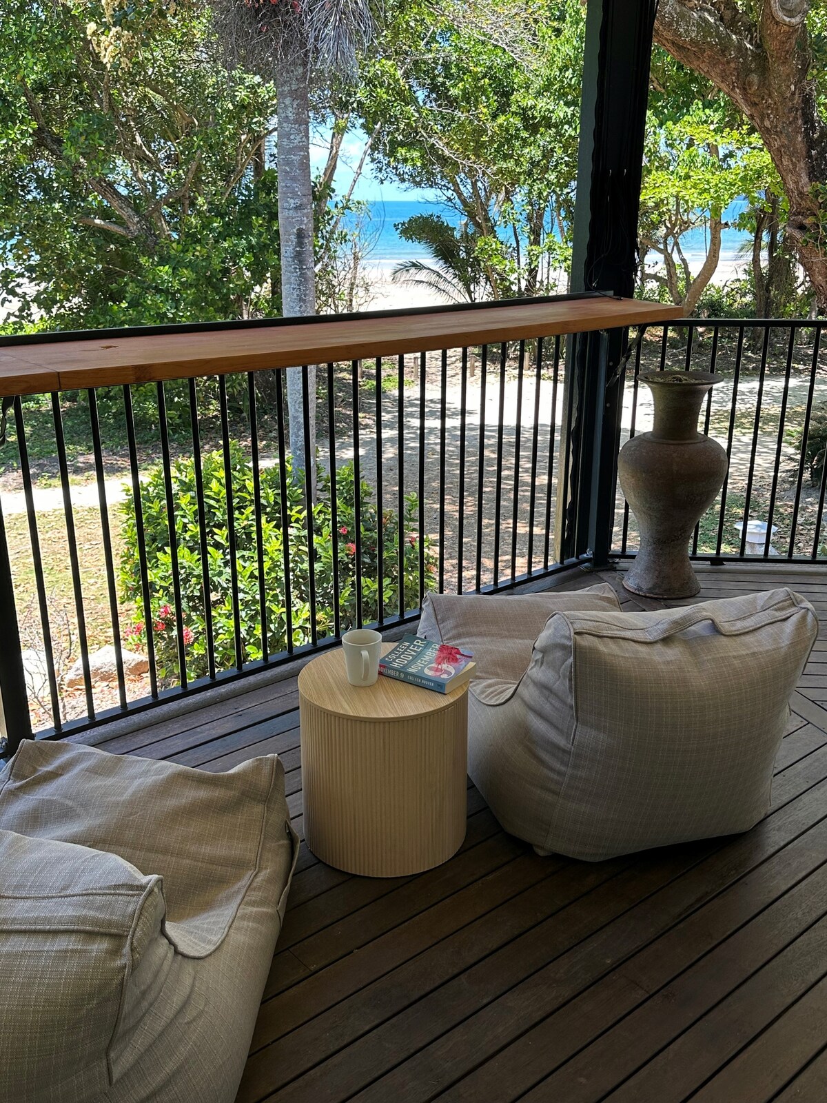 A covered deck space features comfortable bean bag chairs arranged around a small wooden table. A ceramic vase adds a decorative touch. In the background, glimpses of greenery and the beach can be seen through the railing, creating a peaceful outdoor setting.