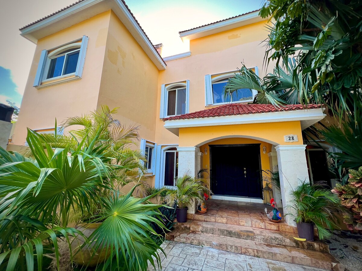 The exterior of the villa is framed by lush greenery, showcasing a vibrant yellow façade. A stone pathway leads to the entrance, which features double doors under a tiled portico. Tropical plants add to the welcoming atmosphere of the home.