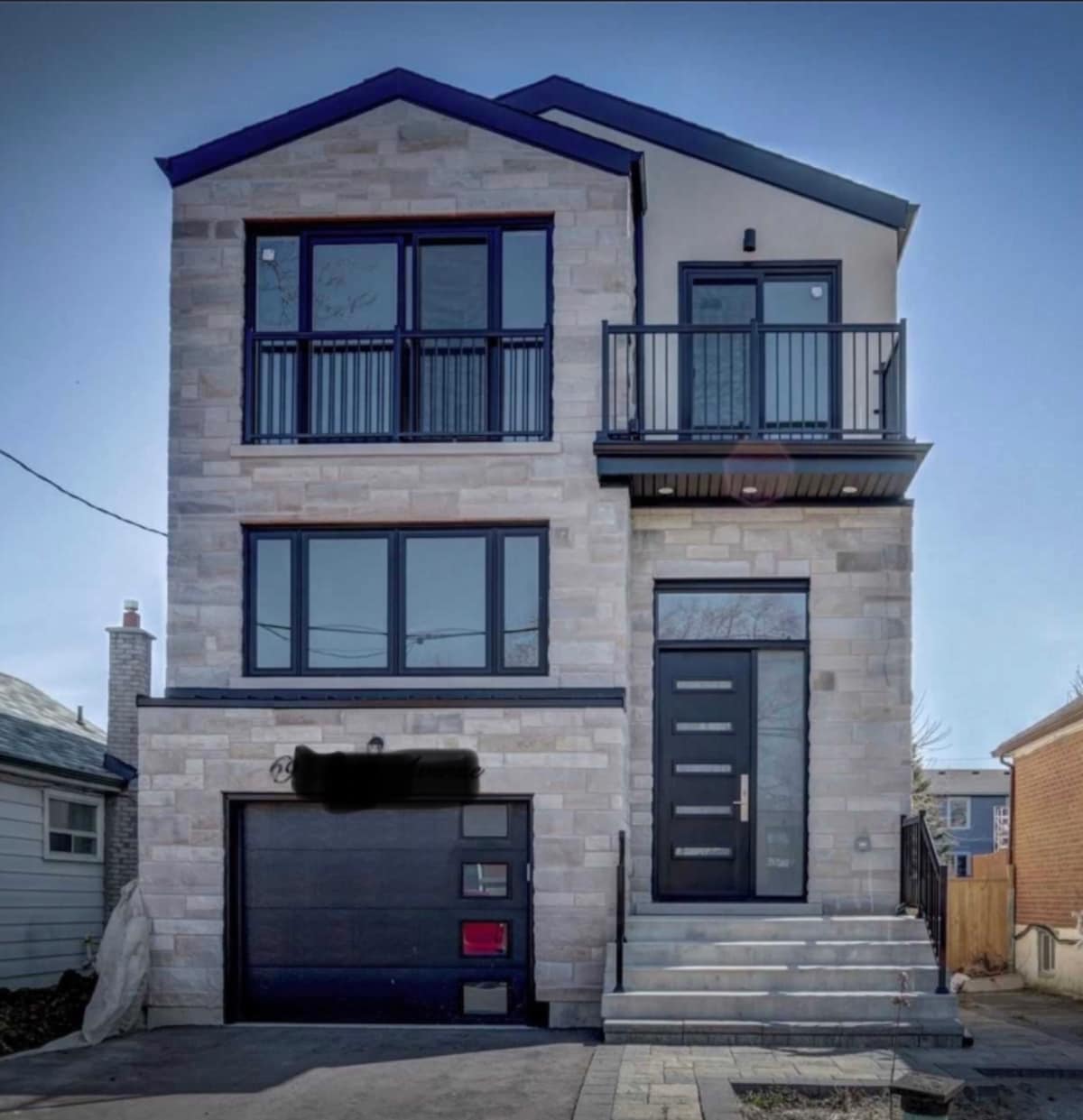 The modern exterior of a newly built three-story home features large, symmetrical windows and a stylish front door. A small balcony extends from the upper floor, and a garage with a sleek design is visible on the ground level. Steps lead to the front entrance.