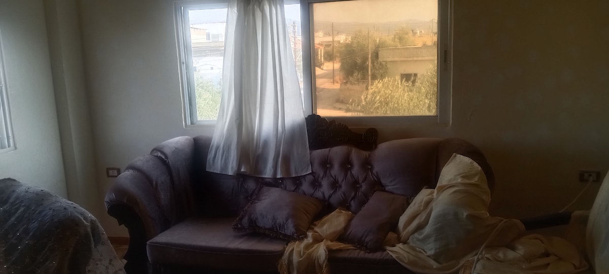 A soft, tufted couch is positioned near large windows that allow natural light to illuminate the room. Light fabric drapes gently over one arm of the couch, while decorative pillows add subtle texture. A glimpse of the outside landscape is visible through the windows.