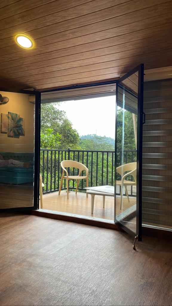 An open glass door leads to a private balcony featuring a small table and two chairs. Lush greenery is visible in the background, enhancing the serene outdoor view.