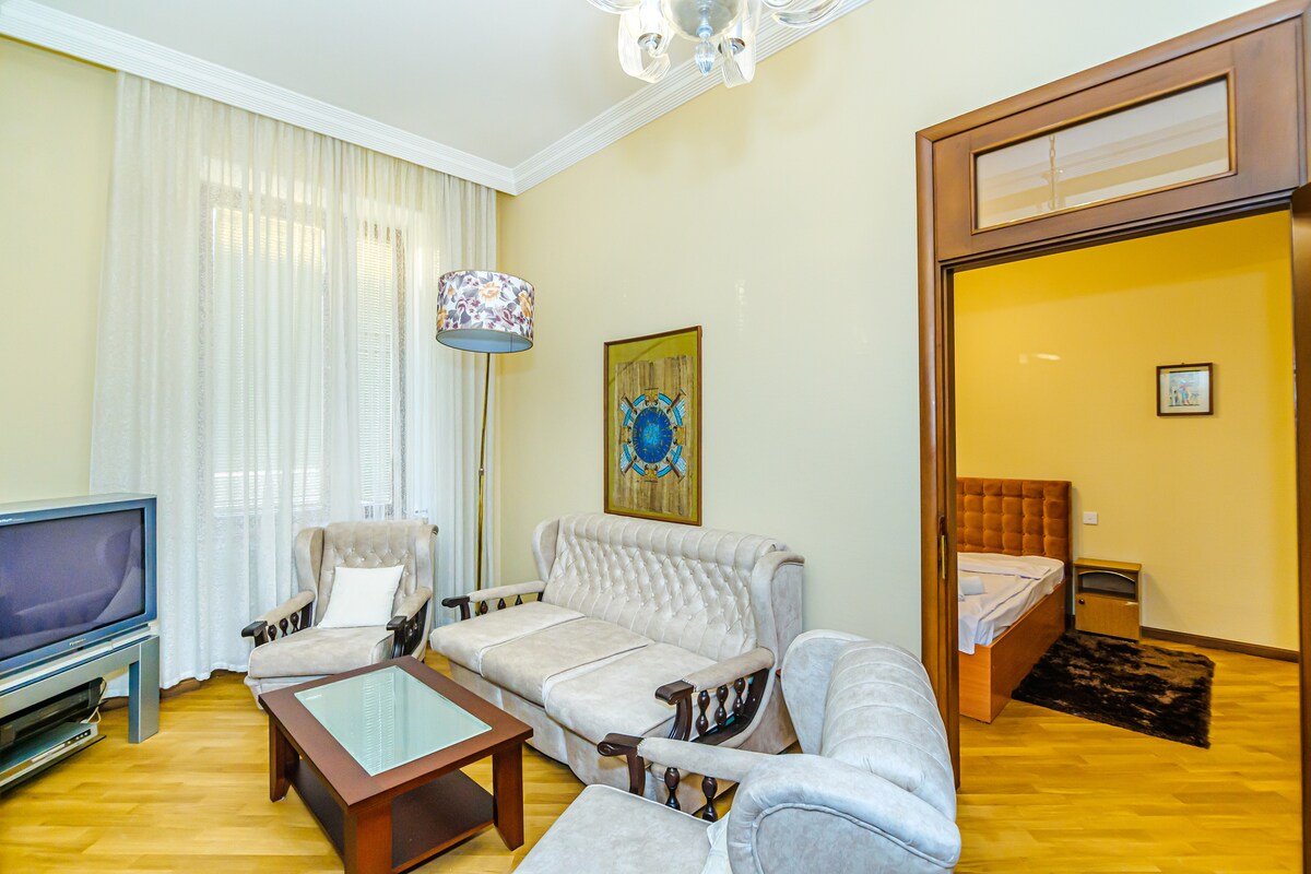 A light-filled living area features a plush seating arrangement with a sofa and armchairs, complemented by a glass coffee table. Sheer curtains allow natural light to fill the space, while a decorative lamp adds warmth. An adjoining bedroom is partially visible through an open doorway.