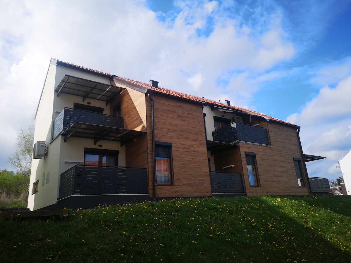 A two-story building is positioned on a grassy incline, featuring a blend of wood and stucco exteriors. Each apartment is equipped with a private balcony. The sky is partly cloudy, creating a serene backdrop for the structure.