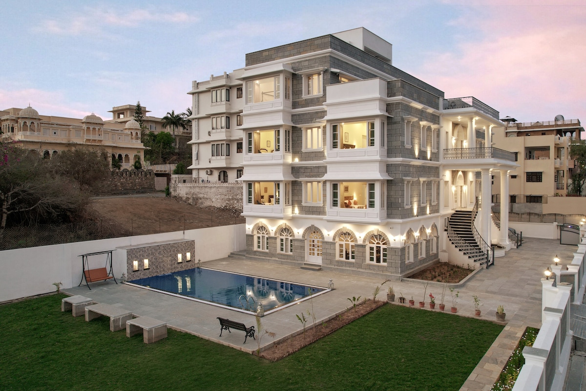 A modern three-story building is shown, featuring numerous windows and balconies. A swimming pool is situated in front, surrounded by a spacious lawn with lounge areas. The structure's facade combines stone and white elements, contributing to its contemporary charm against a softly lit evening sky.
