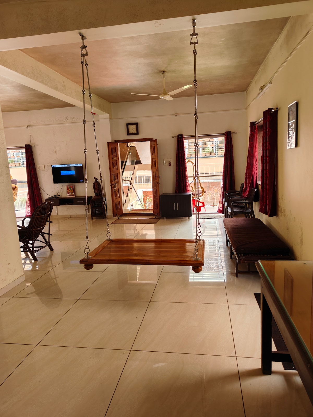 A spacious indoor area features a traditional wooden swing suspended from the ceiling. Comfortable seating arrangements, including a sofa and chairs, are present. Large windows covered by red curtains allow natural light to fill the room, while a television is mounted on the wall.