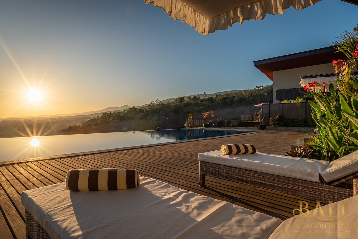 An infinity pool reflects the soft glow of sunrise, with a wooden deck featuring two sun loungers adorned with striped cushions. In the background, lush greenery and distant hills are illuminated by the morning light, enhancing the tranquil atmosphere.