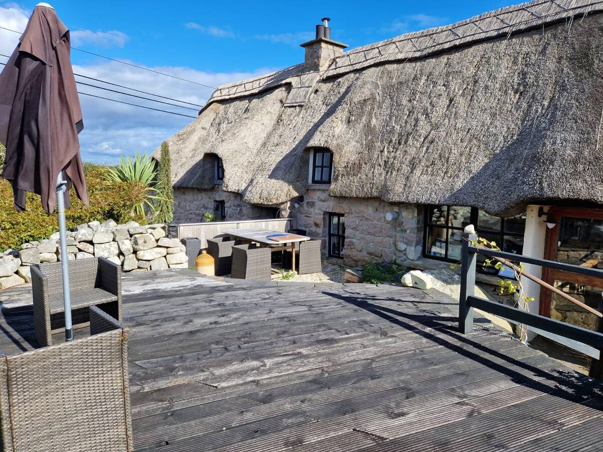 A thatched-roof cottage is showcased, surrounded by a wooden deck. Outdoor seating is provided with a table and chairs, and a patio umbrella is positioned nearby. A backdrop of rustic stone walls and greenery adds to the natural setting.