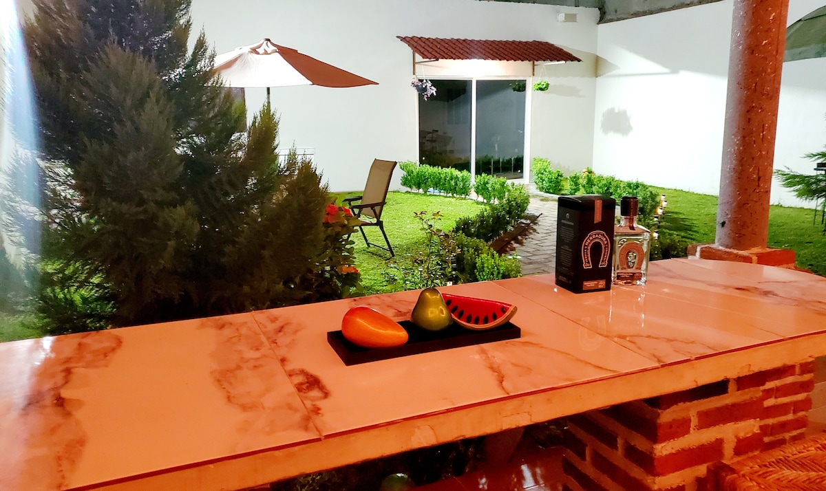 An outdoor space is highlighted, featuring a marble-topped bar with colorful fruit displayed on a black platter. In the background, a well-maintained garden with greenery is visible, along with a lounge chair and a shaded area provided by an umbrella.