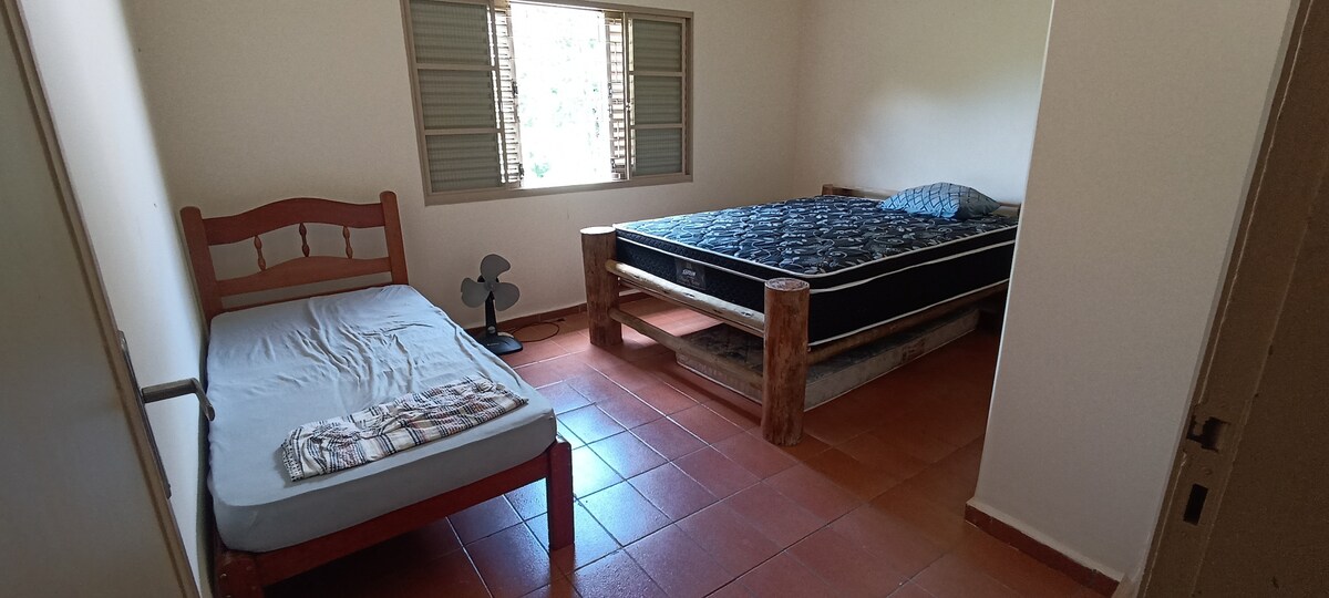A simple bedroom is featured with two beds: a spacious bed with a black mattress and a smaller bed with a light mattress. A ceiling fan stands in the corner, and a window provides natural light, framed by wooden shutters.
