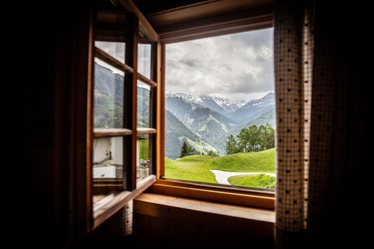 A wooden window is framed with checkered curtains, displaying a panoramic view of rolling green hills and snow-capped mountains. The landscape reveals an inviting path meandering through nature, enhancing the sense of tranquility and connection to the outdoors.