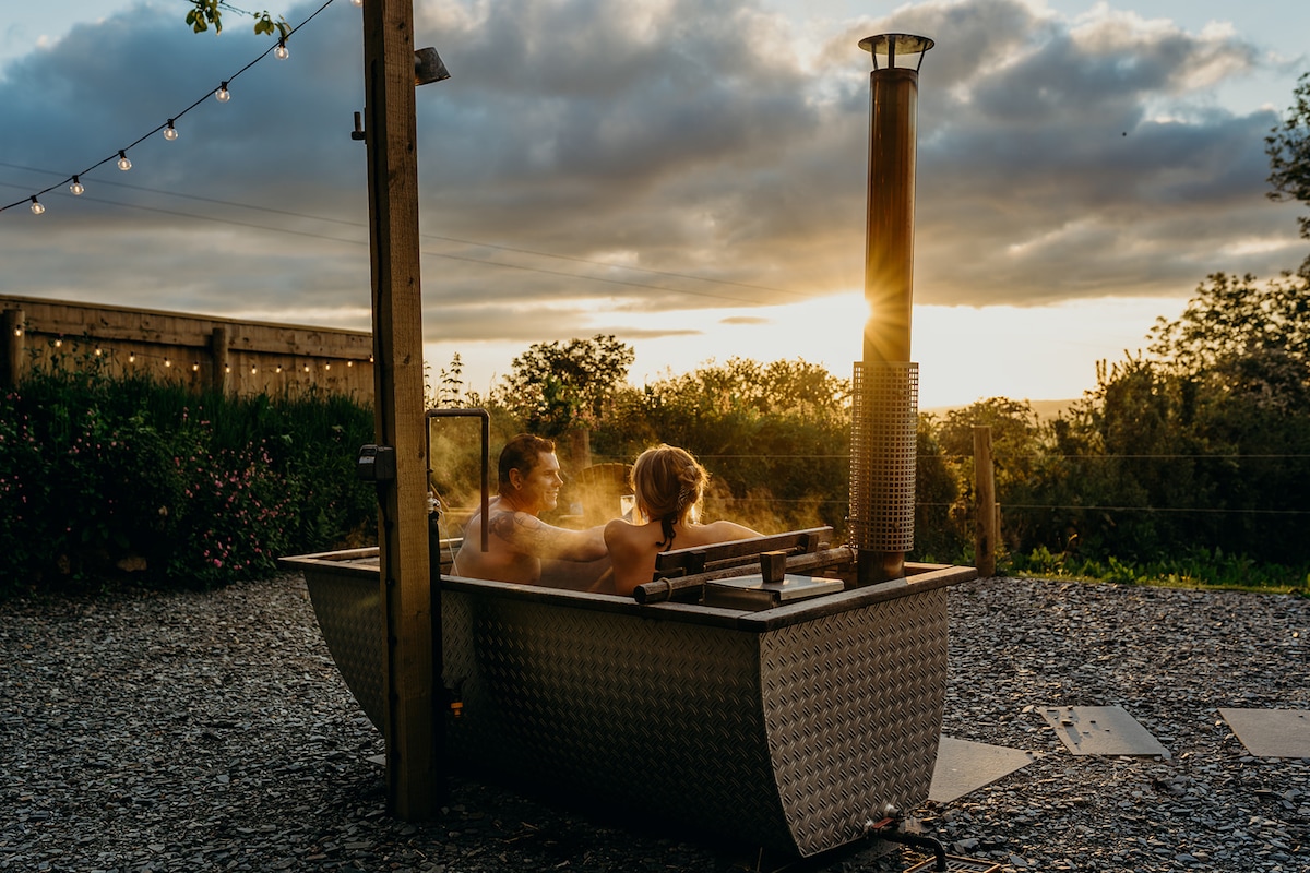 A private hot tub is situated outdoors, surrounded by a gravel patio, where steam rises into the evening air. The sun sets in the background, casting a warm glow over the scenic landscape, enhancing the tranquility of the serene atmosphere.