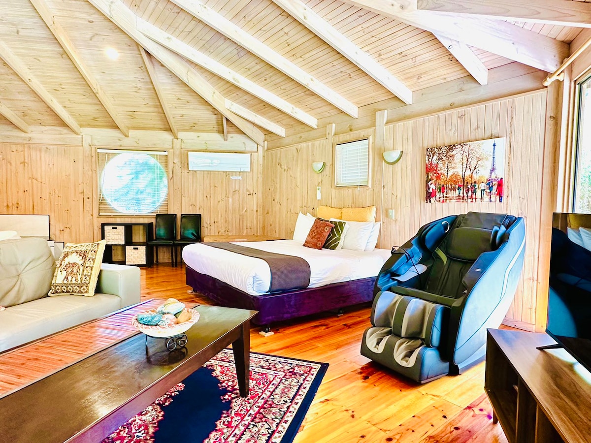 The open-plan timber room features a spacious king bed with soft linens and comfortable seating, including a massage chair. A dark wooden table sits on a patterned rug, while light floods in through large windows, illuminating the natural wood walls and ceiling.