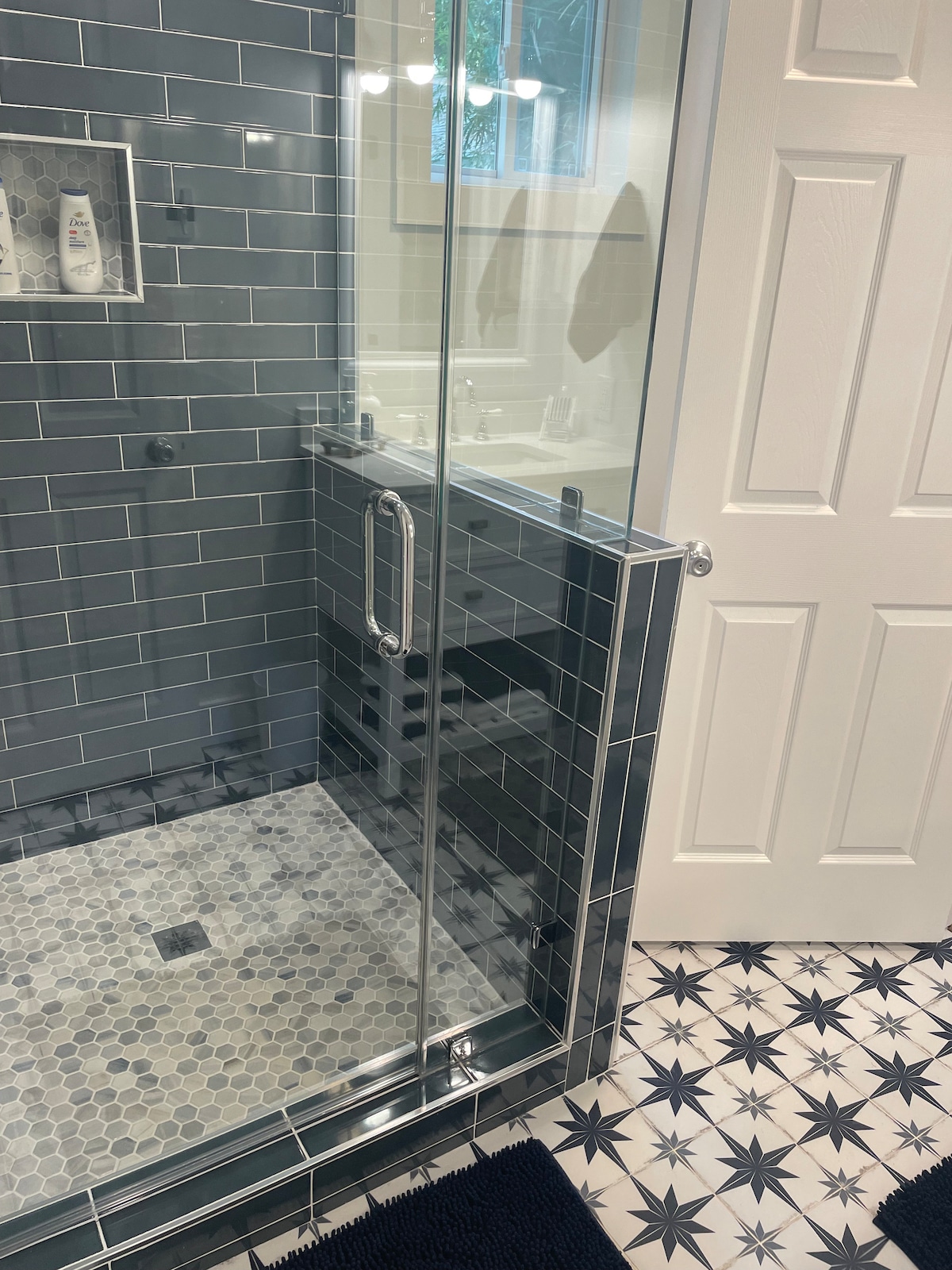 A stylish bathroom features a glass-enclosed shower with dark tile and a hexagonal patterned floor. The space is complemented by a clean white door and a glimpse of a well-lit vanity in the background. Soft blue mats add a touch of comfort.
