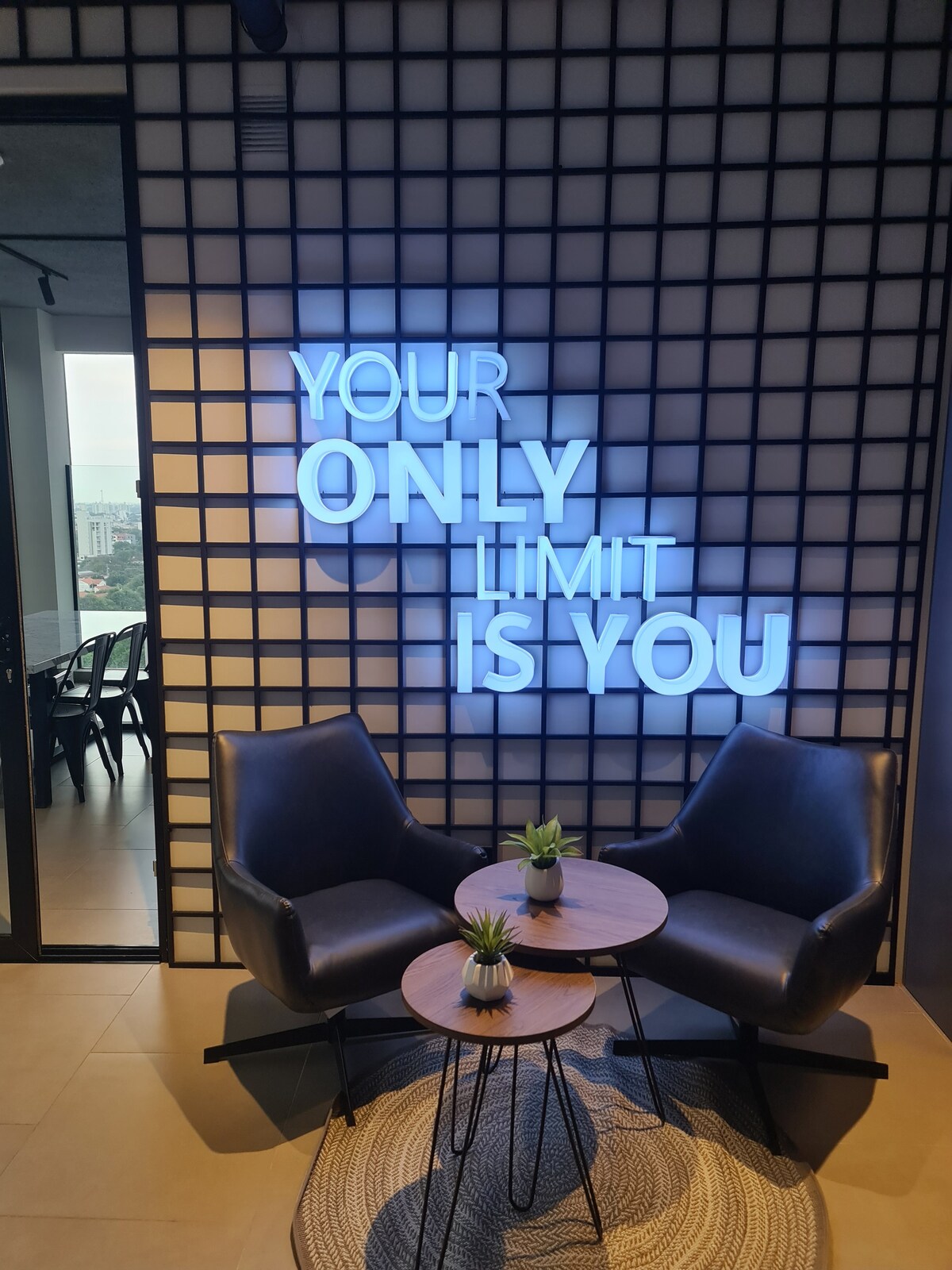 A cozy seating area is highlighted by two dark chairs placed beside a small table. A decorative plant sits on the table, while a prominent neon sign conveys a motivational message. The space features a grid-patterned wall and large windows in the background.