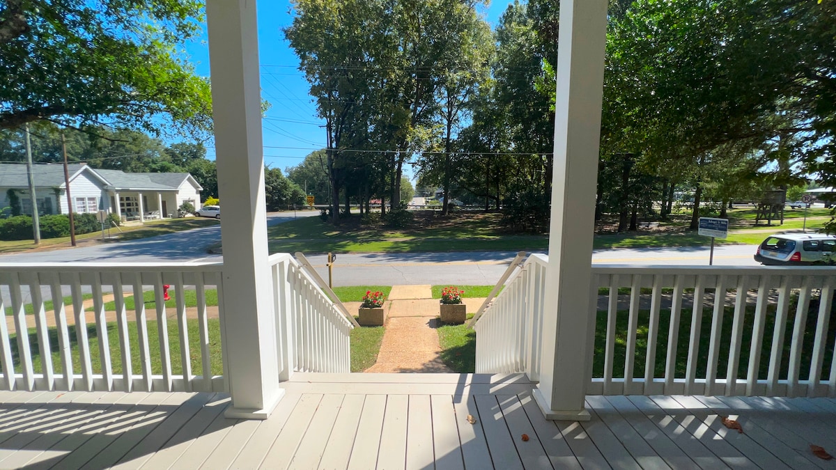 A view from the covered front porch reveals a clear street lined with trees. Flower planters are positioned on either side of the steps, enhancing the entrance. The surrounding greenery offers a peaceful ambiance, inviting guests to enjoy outdoor moments.