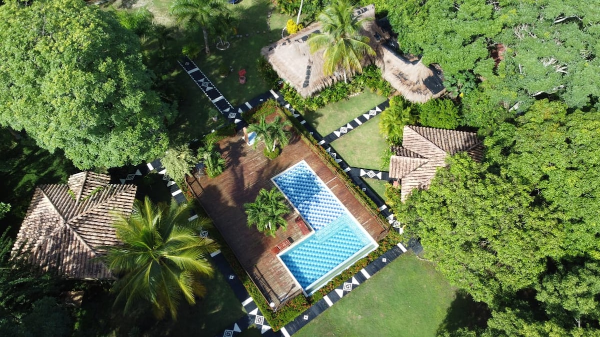An aerial view of a lush outdoor area featuring two distinct swimming pools surrounded by wooden decking. The space is encompassed by tropical greenery, with thatched-roof seating areas and walkways leading through the landscaped garden.