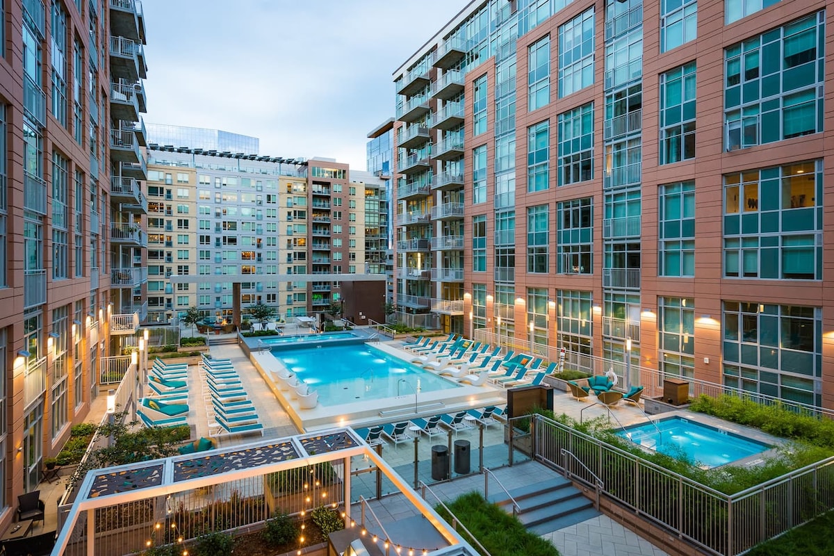 A spacious outdoor pool area is surrounded by modern buildings, featuring numerous lounge chairs along the water's edge. The well-lit space includes two pools, positioned under a clear sky, providing a relaxing environment for guests.
