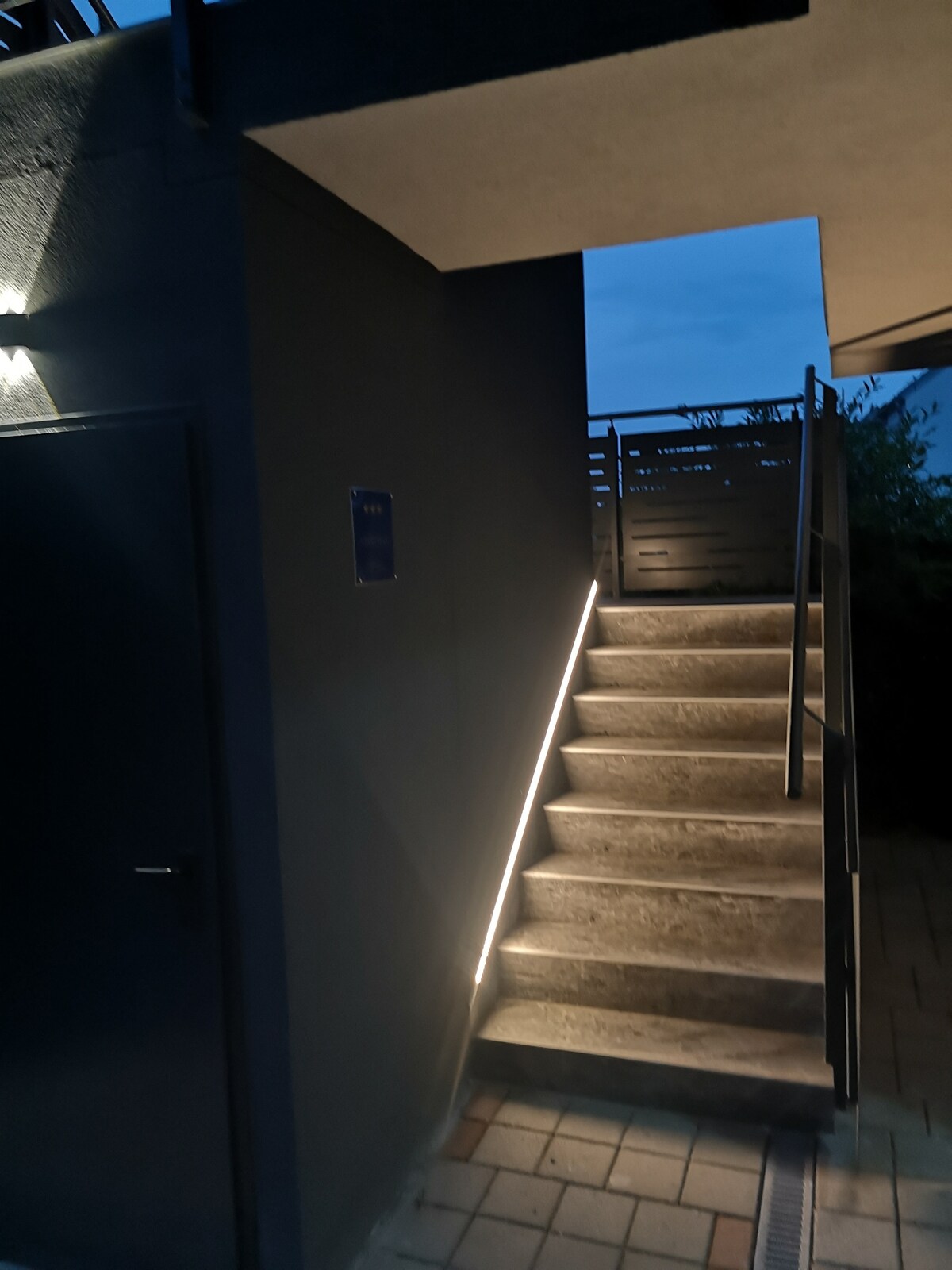 A well-lit staircase is featured, leading upward with soft illumination along the sides. The stairs are positioned next to a dark wall, enhancing the modern design of the outdoor space. A railing is visible, providing safety as individuals ascend.