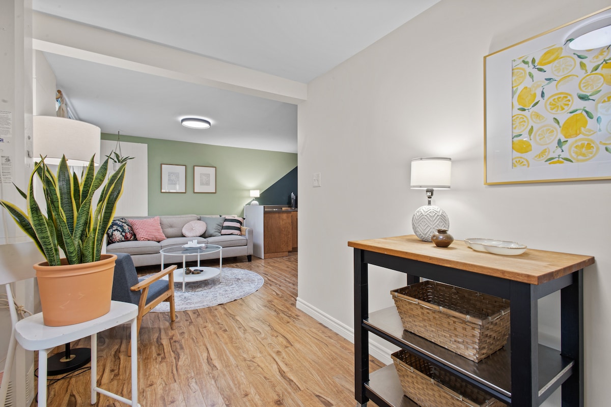 An open concept living area features a cozy sofa, round coffee table, and warm wood flooring. A potted plant adds a touch of greenery. A small side table with a lamp provides ambient lighting, while a colorful art piece complements the decor.
