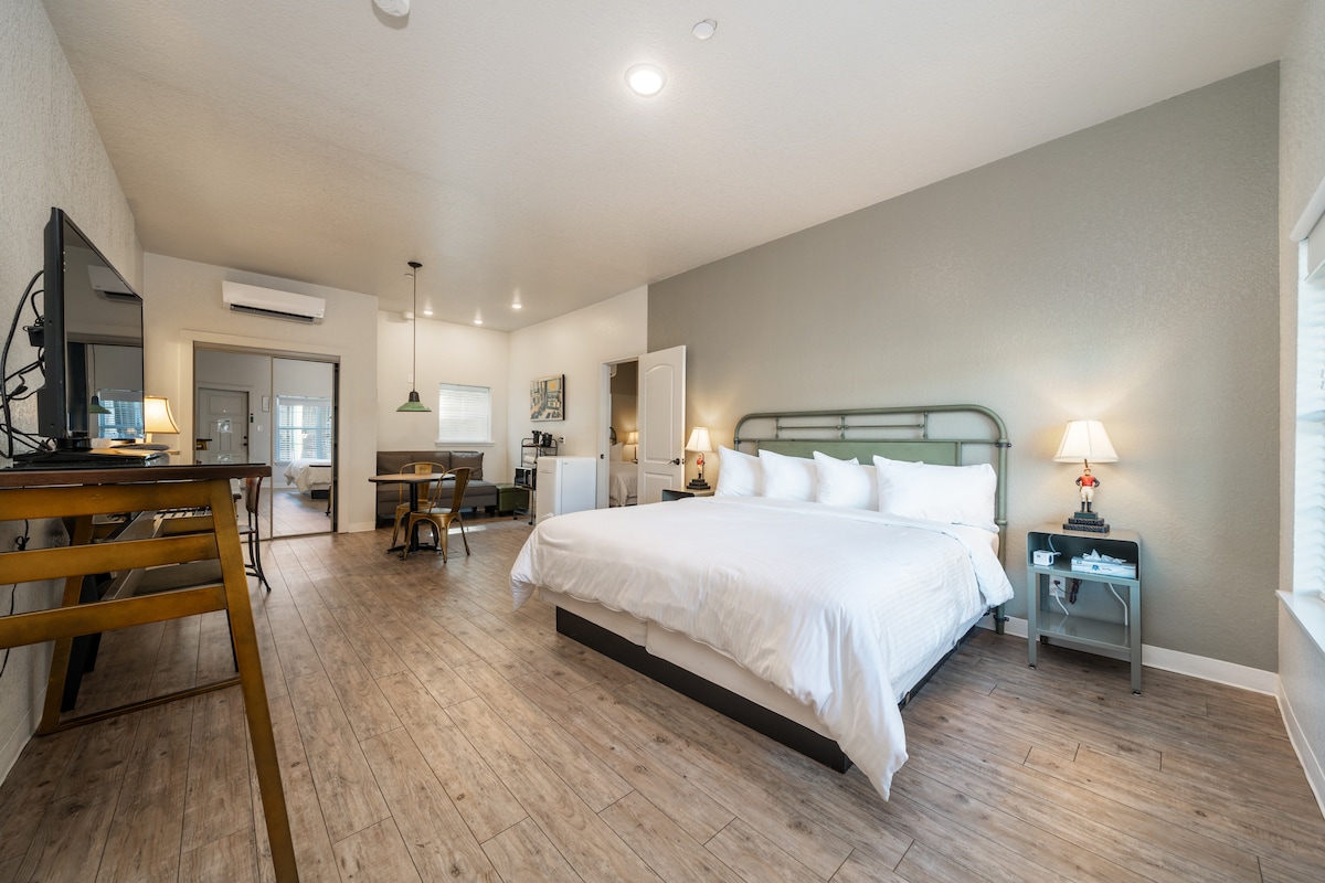 A spacious studio-style suite features a king bed with white linens, accompanied by bedside lamps. A work desk and dining table for four are positioned across the room. A flat-screen television is mounted on the wall, with large windows allowing natural light inside.