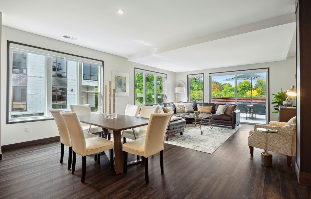 A spacious living area features large windows that invite natural light, displaying views of greenery outside. A dining table is set for six, while a cozy seating arrangement includes a dark leather sofa and an armchair, accented by a decorative area rug.