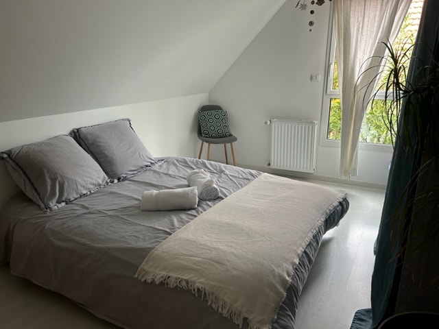 A private bedroom features a spacious bed covered with neutral-toned linens and a soft throw. A small chair with a patterned cushion stands nearby, while natural light enters through a window adorned with light curtains. A towel is neatly placed on the bed.