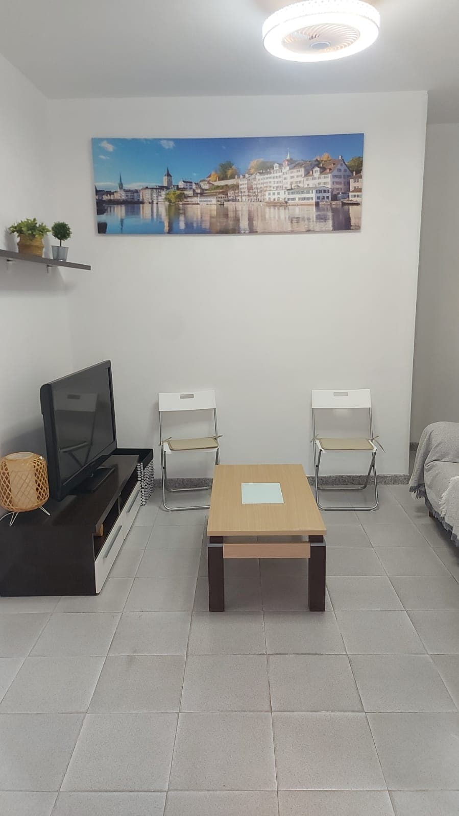 A living area is presented with a sleek television mounted on a dark, low-profile cabinet. A light-colored coffee table is centered between two white folding chairs. A scenic wall art featuring a waterfront view hangs above the seating area, and a potted plant adds a touch of greenery.