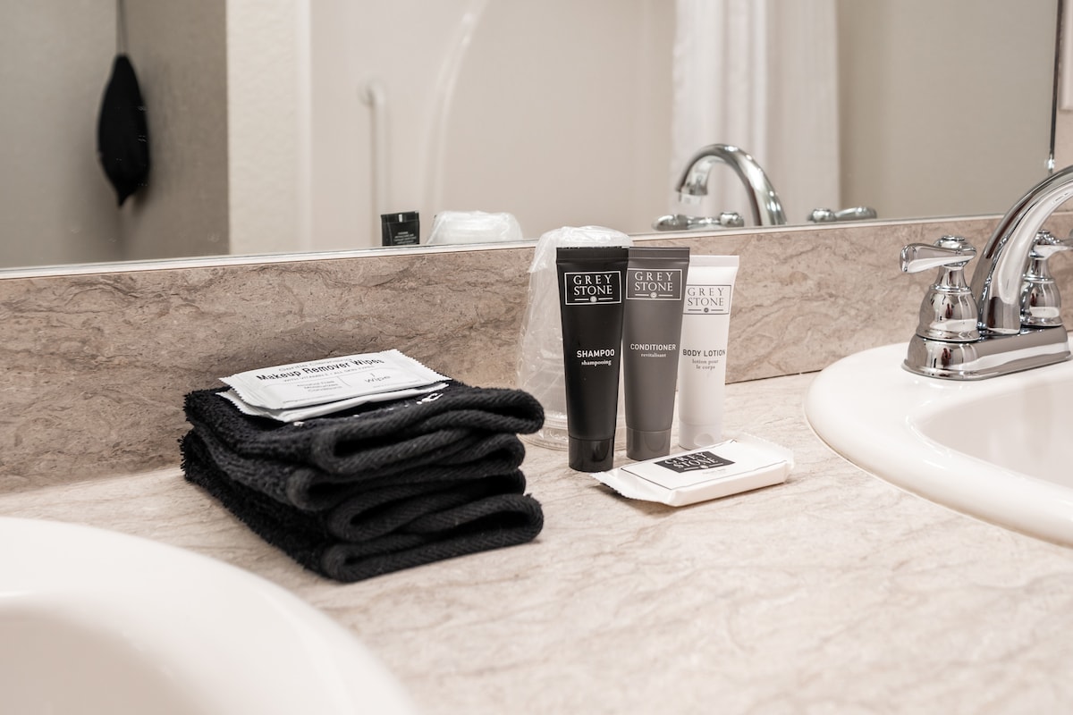 A well-arranged bathroom counter displays complimentary toiletries including shampoo, conditioner, and body lotion in stylish tubes. Dark towels are neatly stacked beside a soap pouch, all set against a light-colored countertop. The mirror reflects a portion of the sink area and soft lighting from the overhead fixtures.