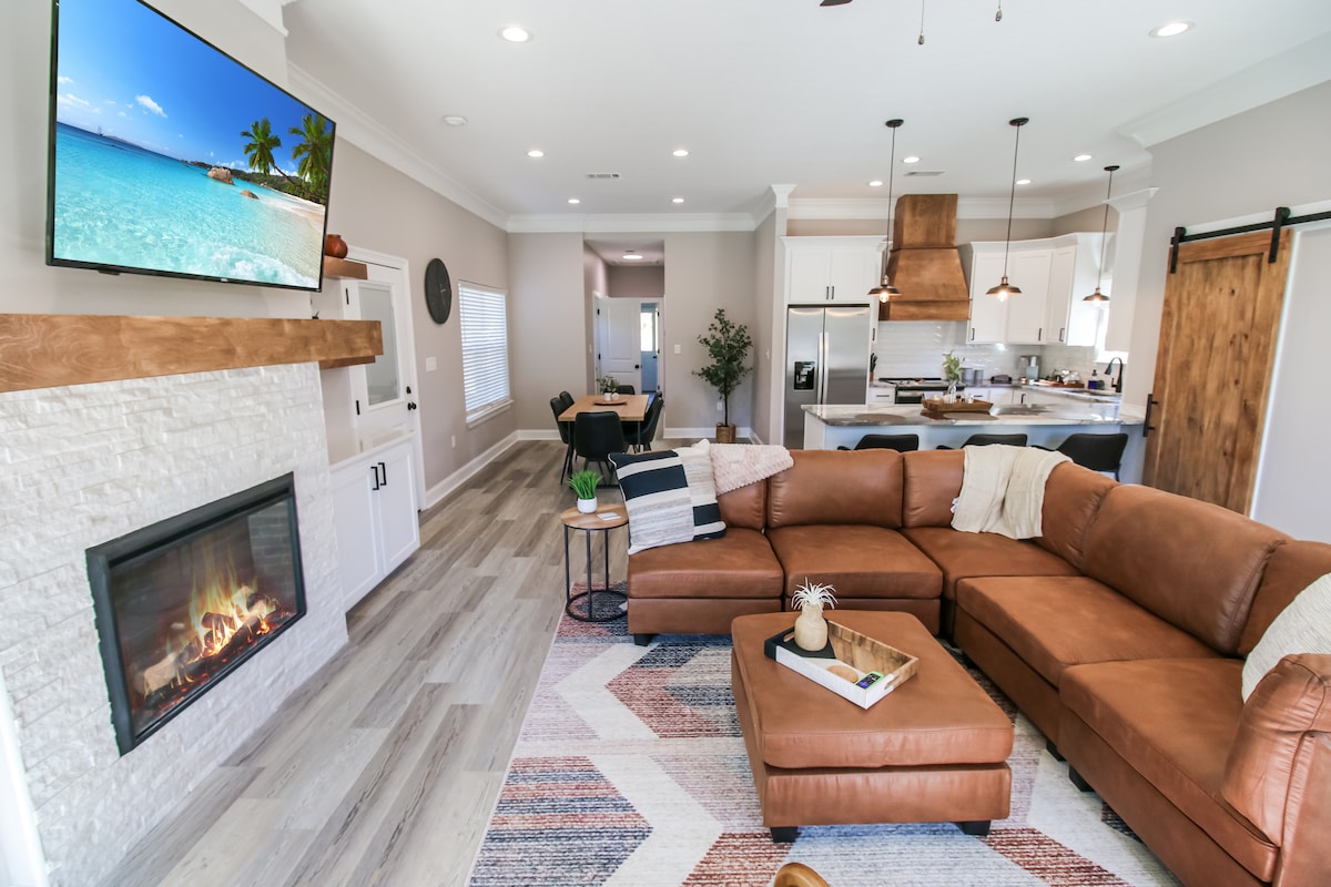 A spacious living area features a comfortable leather sectional sofa arranged around a patterned area rug. A modern fireplace is complemented by a natural wood mantel. A television mounted on the wall displays a tropical beach scene, enhancing the coastal ambiance.