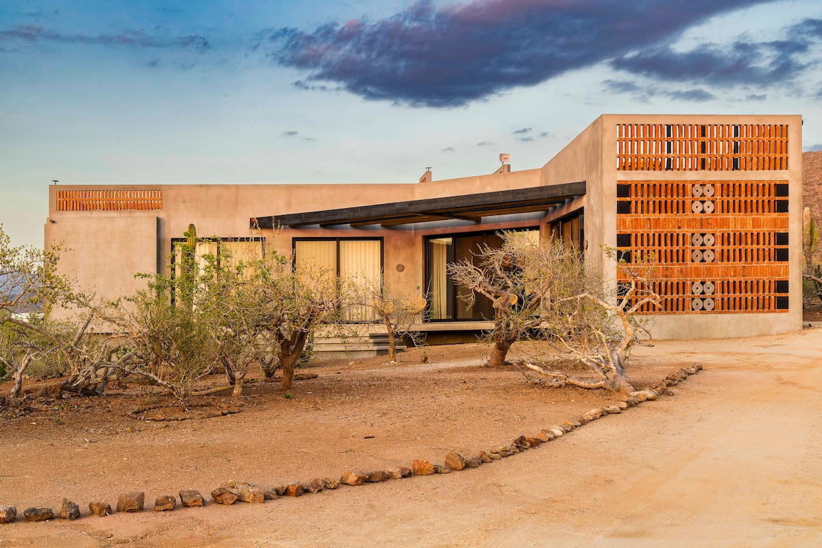 A modern structure with earthy tones and intricate wooden screens is complemented by natural landscaping. The design incorporates large windows that allow ample light. The approach is marked by a gravel path lined with low vegetation, enhancing the serene desert environment.
