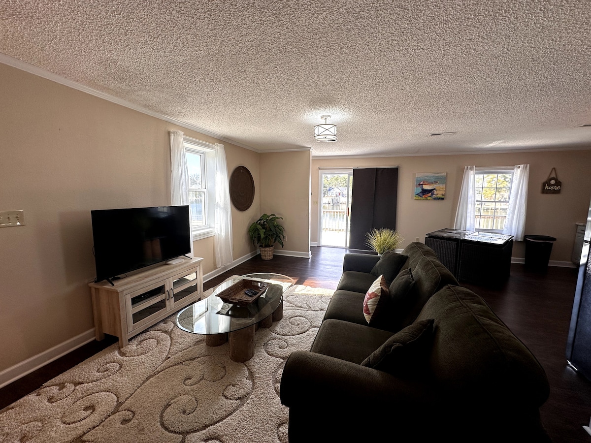The living area is comfortably arranged with a dark green couch facing a television on a light wooden stand. A decorative glass table sits in front of the couch, surrounded by a cozy area rug. Sunlight filters through two windows adorned with sheer curtains.