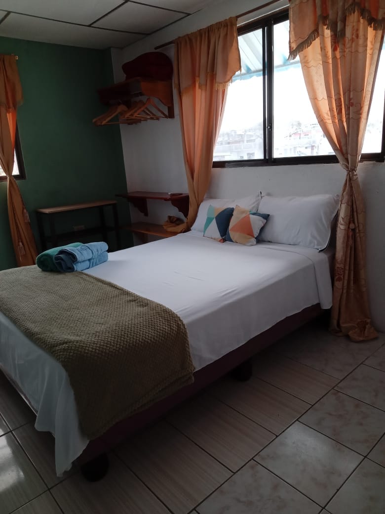 A cozy bedroom features a spacious bed dressed in white linens, complemented by colorful accent pillows. Natural light filters through two windows, adorned with light curtains. A small table and wooden shelving provide practical storage options within the warm atmosphere of the room.