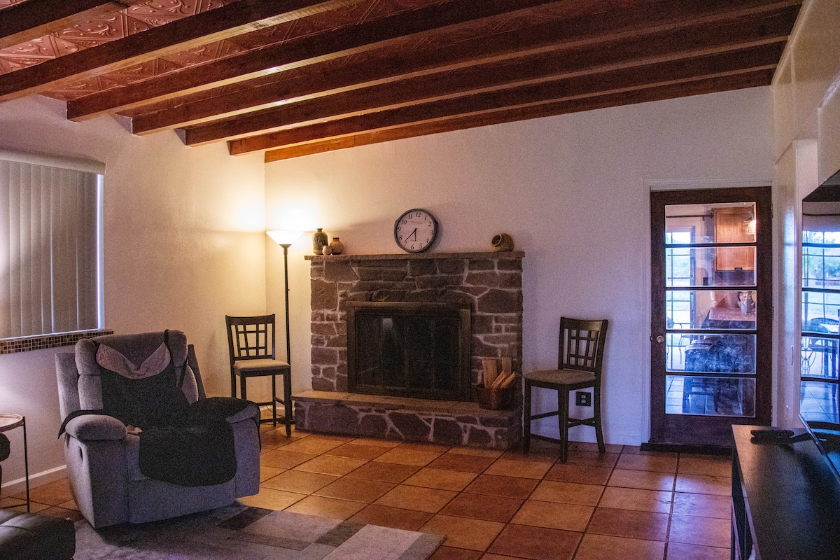 A living area features a stone fireplace as the centerpiece. Two wooden chairs are positioned near the fireplace, while a tall lamp provides soft lighting. Exposed wooden beams enhance the ceiling, and a glass door offers a glimpse into an adjoining room.