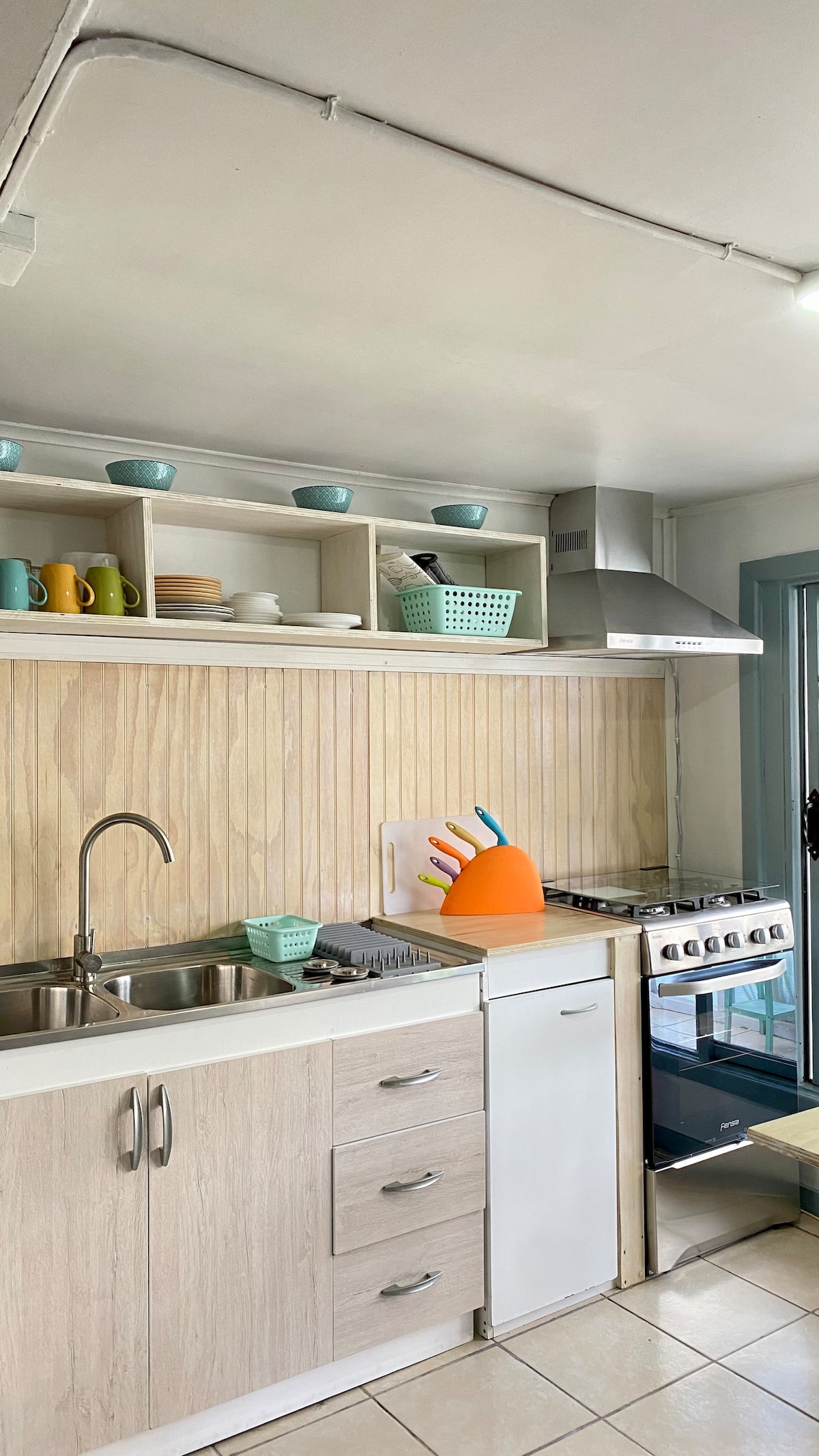 The kitchen features a sleek layout with a stainless steel sink and a gas stove. Shelves above display colorful mugs and plates, while a bright orange utensil holder adds a pop of color. Ample counter space enhances functionality, complemented by tiled flooring.