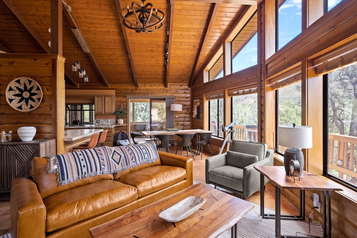 A spacious living area is highlighted by large windows allowing natural light to fill the room. A leather sofa sits invitingly, paired with a green accent chair. The wood-paneled walls and ceiling create a warm atmosphere, with a wooden dining table and modern chairs visible in the background.