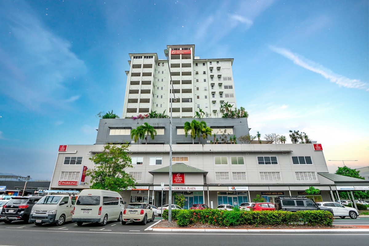 The exterior of the Cairns Central Plaza Apartments complex is presented, highlighting a modern multi-story building with multiple balconies and greenery. The ground level features a commercial area flanked by vehicles parked along the street, showcasing the building's central location.