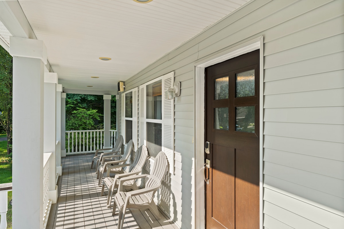 A welcoming porch area features several comfortable chairs arranged on a spacious deck. Natural light filters through the railing, creating patterns on the floor, while greenery is visible in the background, enhancing the outdoor experience.