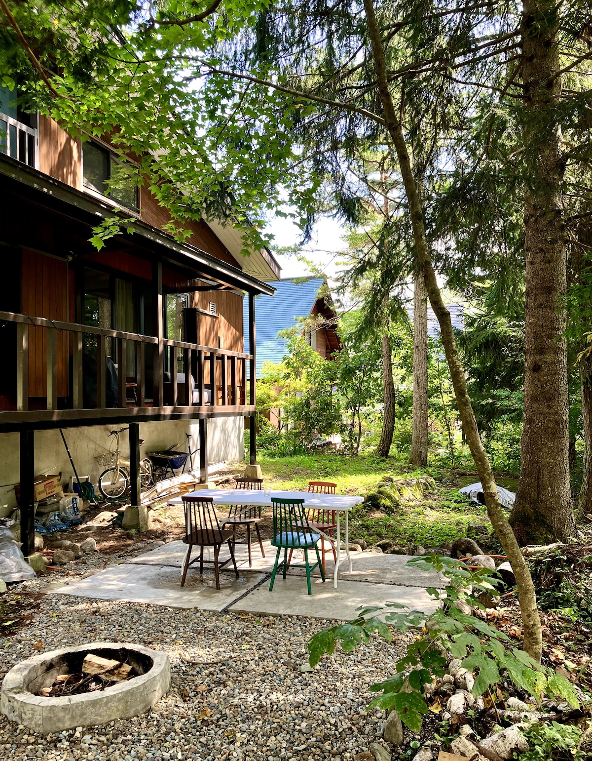 A private backyard area features a stone patio with a table and chairs surrounded by lush greenery. Nearby, a fire pit made of stone is visible, nestled among the trees, offering a peaceful setting for outdoor gatherings.