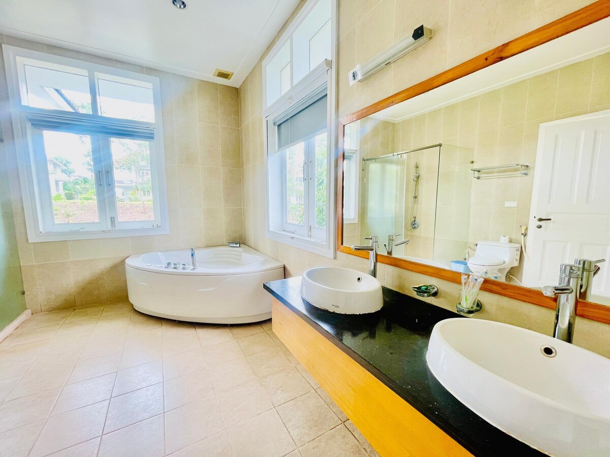 A well-lit bathroom features a spacious soaking tub alongside a sleek wooden countertop with dual sinks. Natural light enters through large windows, illuminating the neutral-toned tiles and creating an open atmosphere. A glass shower area is visible in the corner.