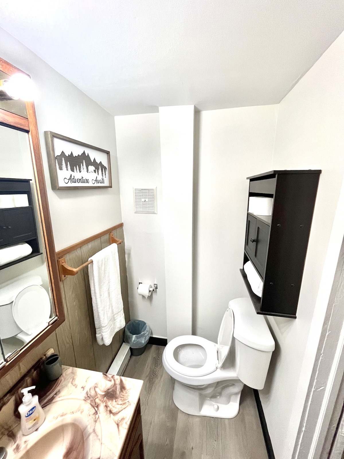 A compact bathroom space features a white toilet and a wall-mounted shelf equipped with towels and toiletries. A large mirror above the sink reflects the room, while a light wood cabinet adds to the decor. A small wastebasket is placed near the toilet.
