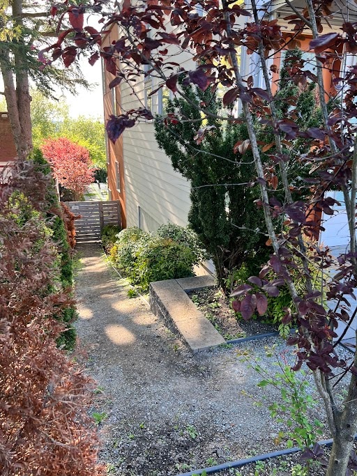 A quiet pathway is framed by lush greenery and ornamental trees. Sunlight filters through the leaves, highlighting a gravel path that leads towards the entrance of the unit. The scene is surrounded by vibrant foliage, creating a calm and inviting outdoor space.