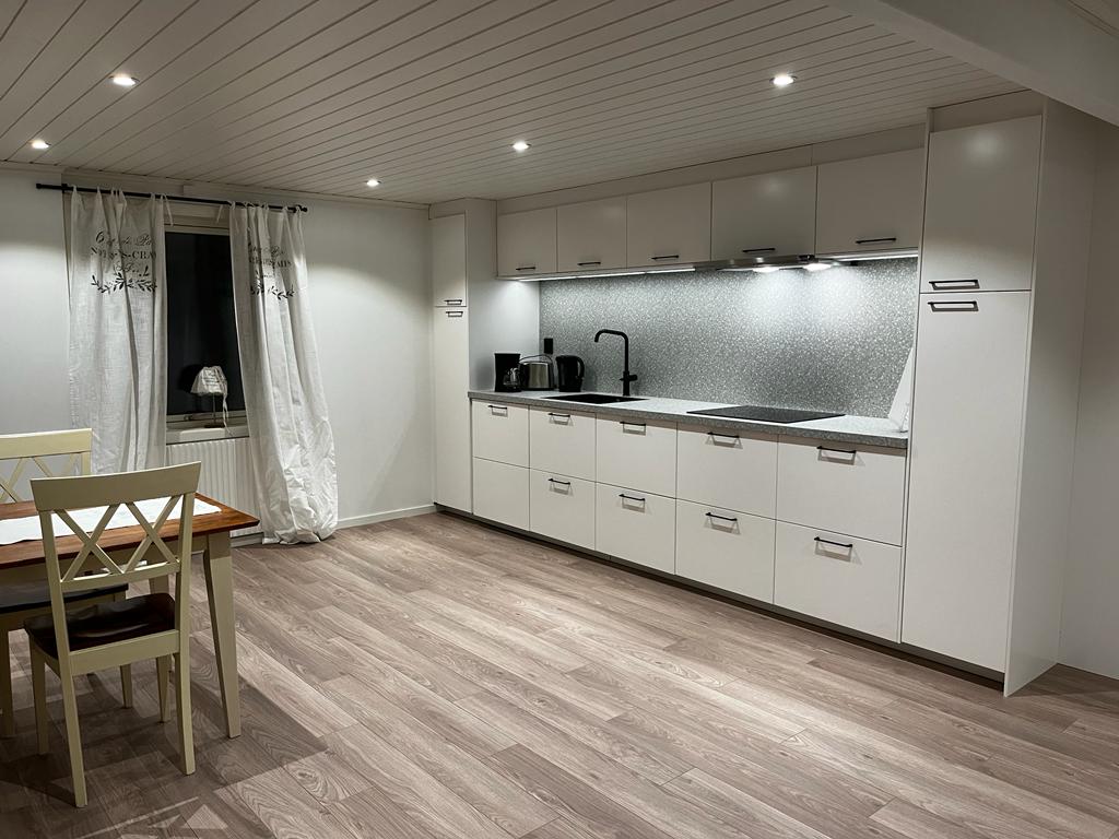 A modern kitchen is presented with sleek white cabinetry and a shimmering backsplash. Task lighting illuminates the countertop, while a dining table with several chairs is positioned against one wall. Neutral-toned flooring extends throughout the space, contributing to a clean and spacious feel.