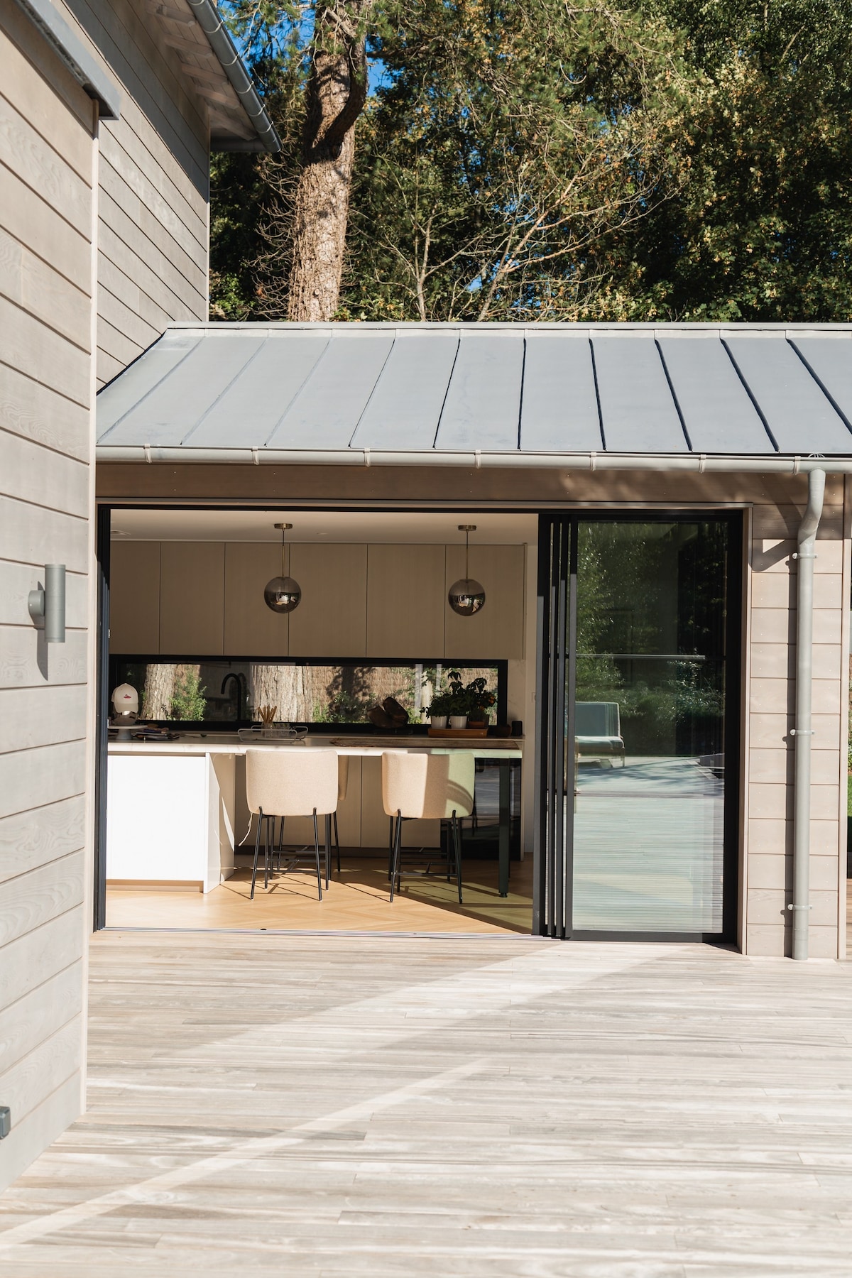 A modern kitchen is visible through large glass doors, featuring sleek cabinetry and minimalistic lighting fixtures. The outdoor wooden deck extends towards the forested area, providing a seamless connection to nature, complemented by a pool visible beyond.