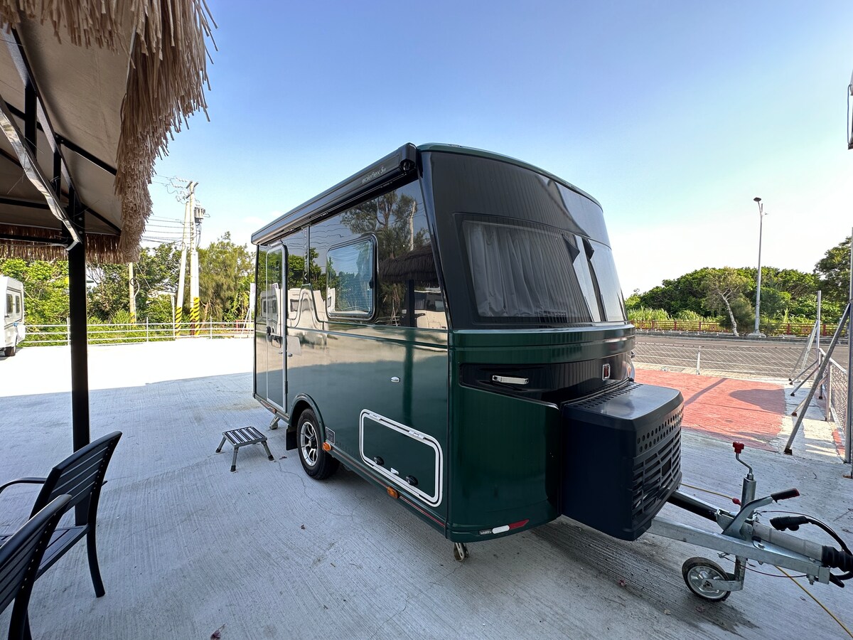 A sleek green camper is parked on a concrete pad, showcasing a modern design. The camper features large windows that provide ample natural light, while the surrounding area includes a shaded seating arrangement made up of black chairs.