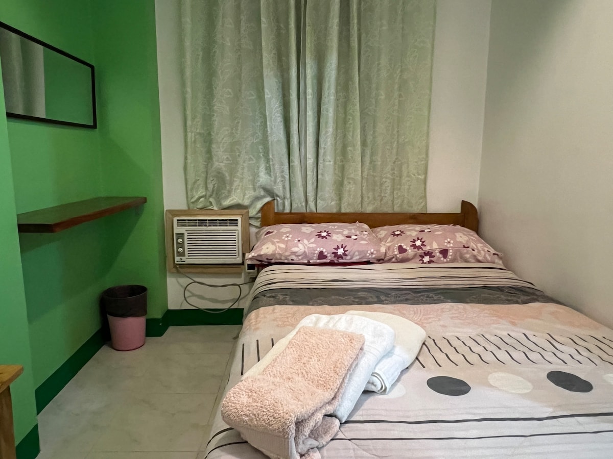 A cozy bedroom features a spacious bed dressed in light bedding with floral accents. An air conditioning unit is mounted on the wall, providing comfort. A small table is visible beside the bed, and soft green curtains frame a window, adding a calm touch to the space.