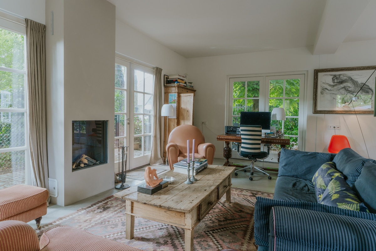 A comfortable living area features a mix of seating options, including a large sofa and two unique armchairs. A wooden coffee table rests on a patterned rug, while an electric fireplace and a stylish desk enhance the space. Natural light frames the room through multiple windows.