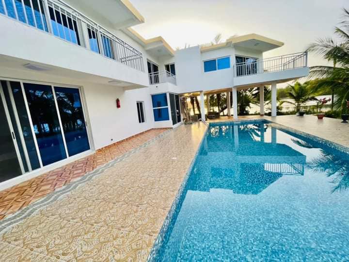 A spacious pool area is surrounded by a modern structure featuring multiple glass doors. The clear water of the pool reflects the serene surroundings, including palm trees. The outdoor space includes a decorative floor patterned in intricate designs leading to the poolside.